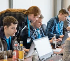 Eine Gruppe von Entwicklern arbeitet gemeinsam an einem Tisch, konzentriert auf ihre Laptops. Eine Person beugt sich vor, um einem anderen Entwickler zu helfen.
