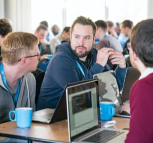 Eine Gruppe von Entwicklern sitzt an einem Tisch und unterhält sich mit Laptops vor sich.
