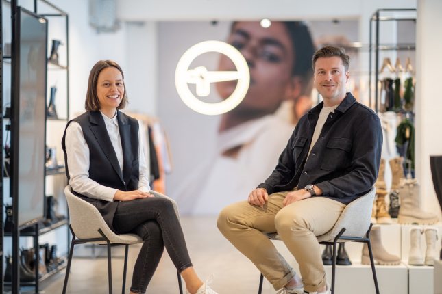 Ein Mann und eine Frau sitzen auf Stühlen in einem modernen Geschäft. Hinter ihnen ist das Tamaris-Logo zu erkennen. Im Hintergrund sind Regale mit verschiedenen Produkten.
